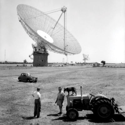 Black and white photo showing a large radio telescope and another in the distance, circular dish-shaped with three long separate arms reaching up from inside the dish and joining above the centre at their top. In the foreground of the treeless flat paddock, two men are talking near a tractor with a dog standing on the tractor.